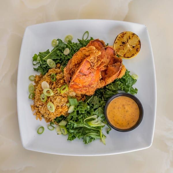 Fried lobster tail with rice, kale, grilled lemon, and a side of sauce on a white plate.