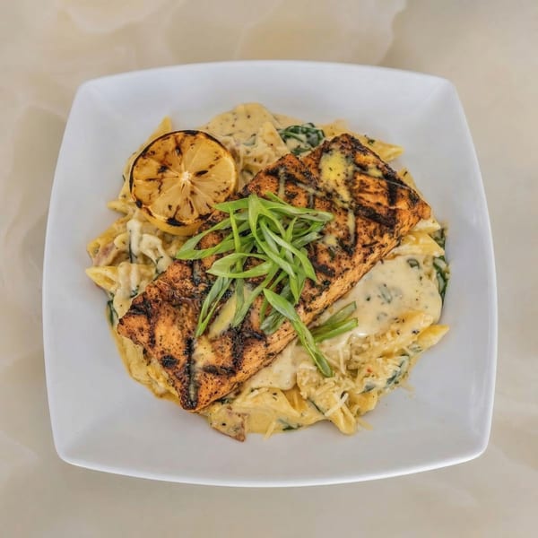 Grilled salmon with green onions and a lemon slice, served over creamy pasta on a white plate.